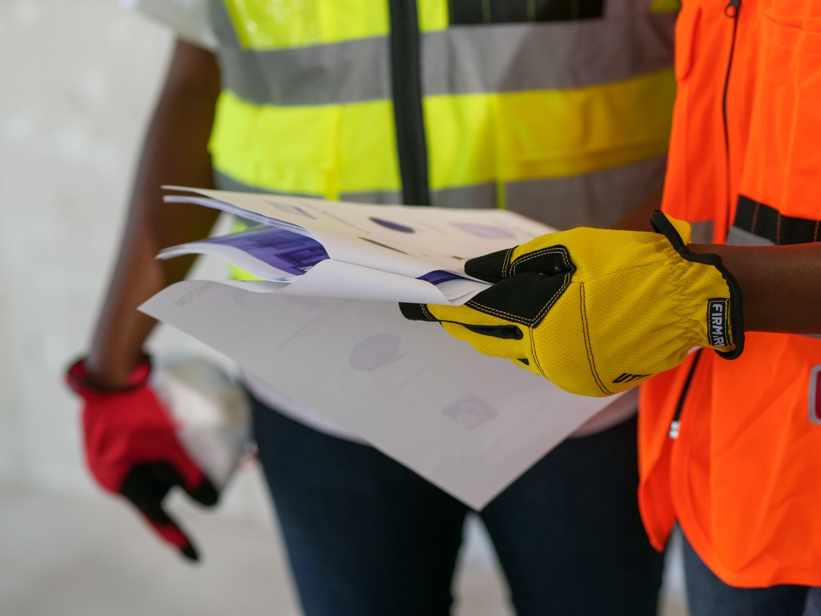 Two workers in hi-vis vests and work gloves reviewing a stack of printed documents on a job site