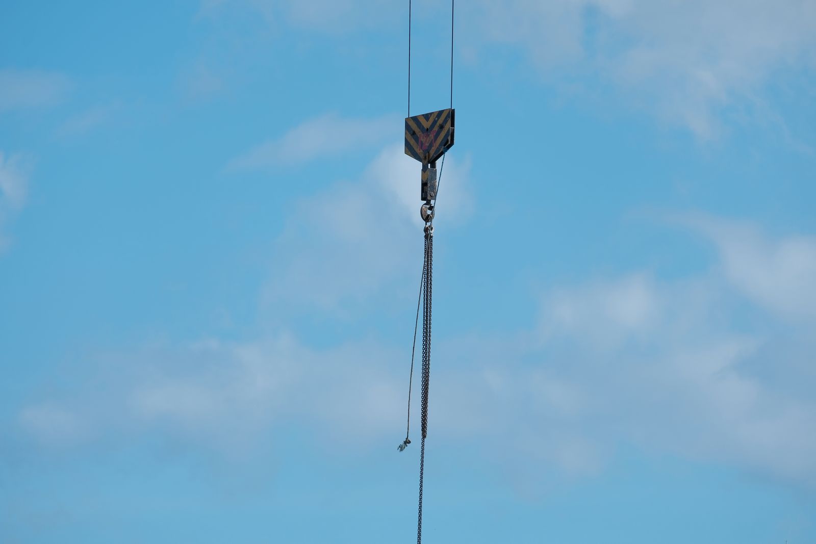 A crane hook block with yellow and black chevron striping hanging from wire rope against a blue sky